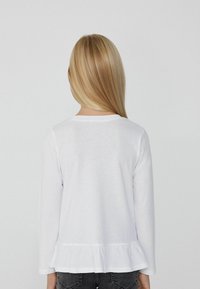 Biała bluzka z długim rękawem i okrągłym dekoltem, z falbaną u dołu, wykonana z miękkiego materiału. Widok z tyłu, z prostymi, blond włosami widocznymi.