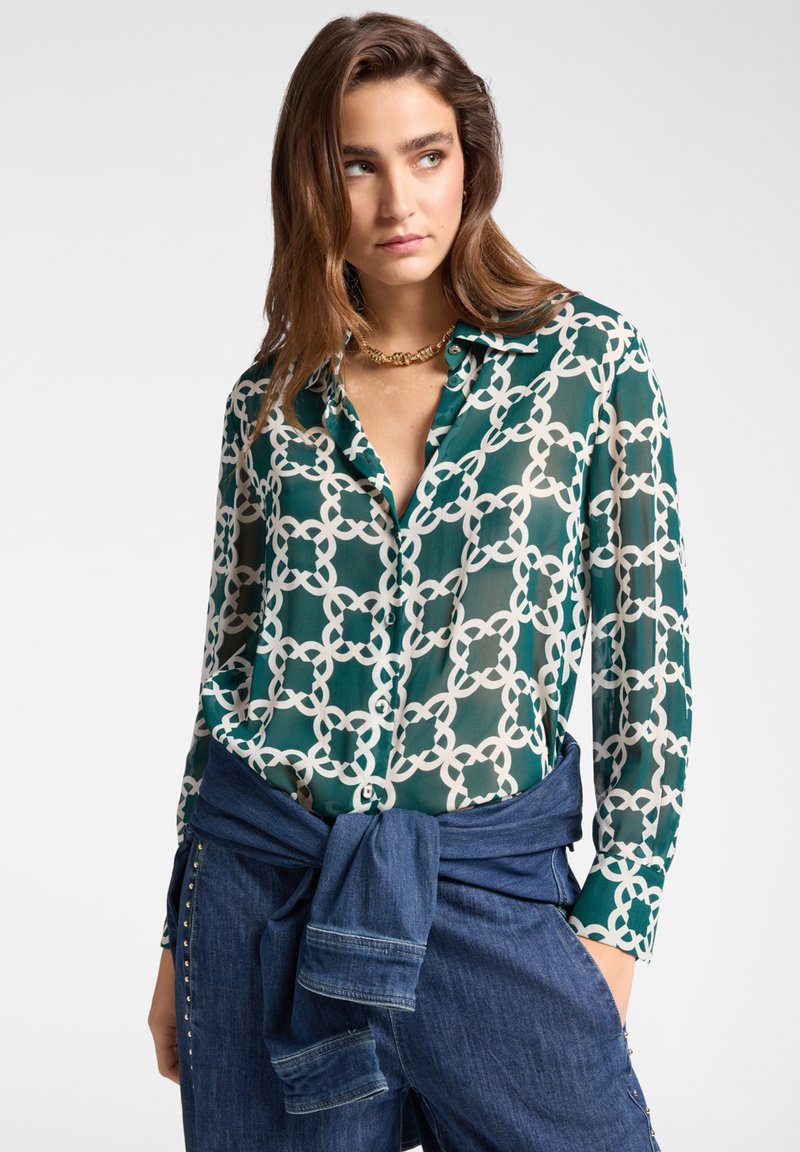 Blusa verde a fantasia con motivo a catena, apertura frontale con bottoni e texture trasparente, abbinata a pantaloni di denim scuri e una cravatta di denim in vita.