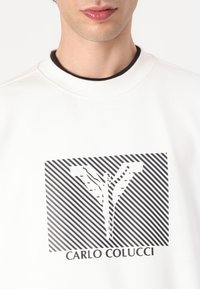 Hvid sweatshirt med en sort grafisk print, der har diagonale striber og en fed "CARLO COLUCCI" tekst nederst. Rund halsudskæring.