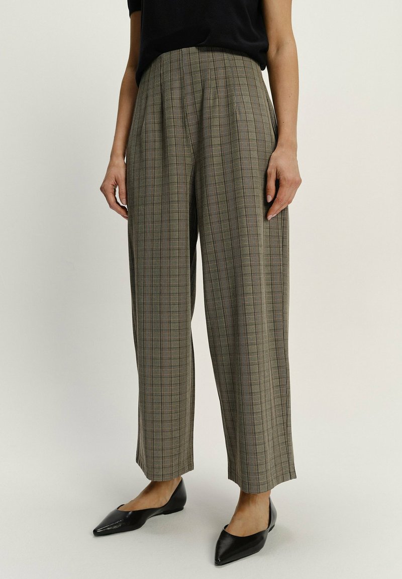 Checked, high-waisted trousers in beige and brown, featuring a loose fit, pleats, and straight legs. Worn with black pointed flats.