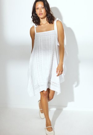 Nainen kävelee valkoisessa hihattomassa mekossa, jossa on pitsiyksityiskohtia, ja valkoiset Mary Jane -kengät valkoista taustaa vasten.