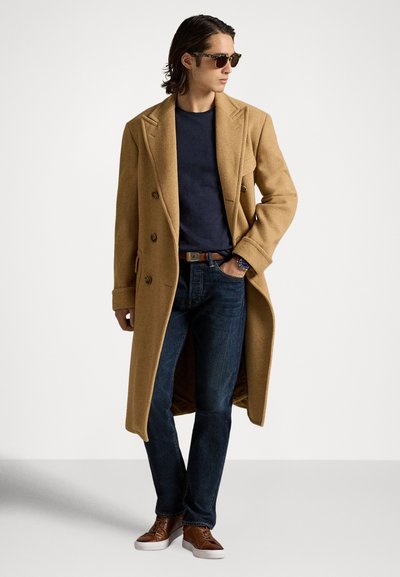 Bézs színű, dupla soros gyapjú kabát, sötétkék póló, sötét farmer, barna sportcipő. Hosszú ujjú és oldalzsebes; napszemüveg egészíti ki a megjelenést.