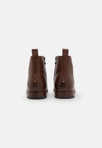 Hnědé kožené kotníkové boty s hladkou texturou, zadním zipovým zapínáním a malou nášivkou s logem poblíž paty. Zaoblená špička a plochá podrážka.