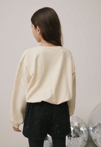 Krémová bavlněná mikina s vyšívanými flitry na rukávech, kombinovaná s černými texturovanými šortkami. V pozadí se nacházejí stříbrné disko koule.