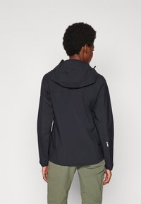 Veste noire imperméable avec capuche, présentant une texture lisse et des poignets ajustables, portée avec un pantalon vert olive clair.