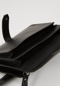 Svart läderplånbok med texturerad yta, tre fack och avtagbar rem. Inkluderar tryckknappsöppningar och subtila sömnadsdetaljer.