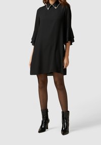 Schwarzes Kleid mit einem kragenförmigen Halsauschnitt, verziert mit Perlenakzenten, weit ausgestellten Dreiviertelärmeln und einer lockeren Passform, kombiniert mit schwarzen Knöchelstiefeln.
