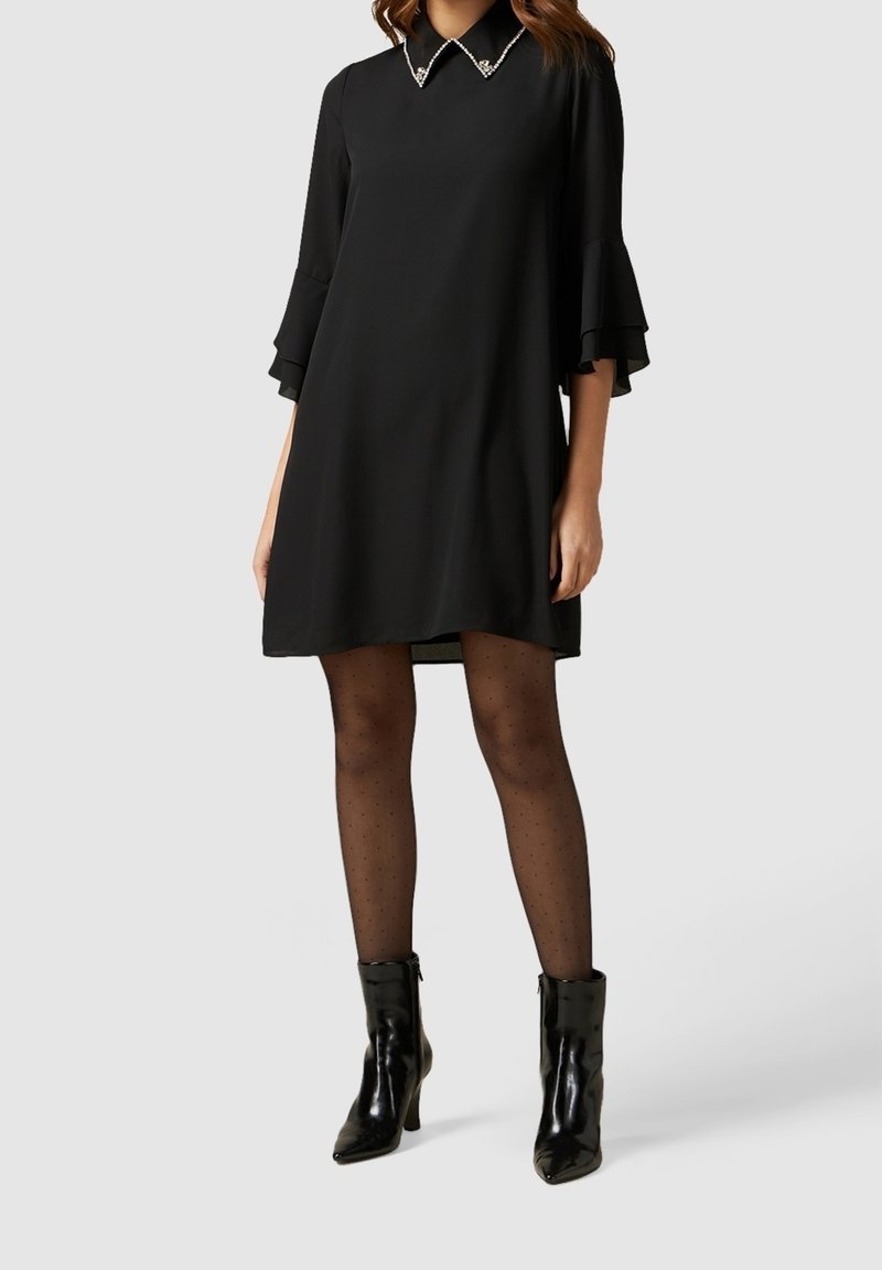 Schwarzes Kleid mit einem kragenförmigen Halsauschnitt, verziert mit Perlenakzenten, weit ausgestellten Dreiviertelärmeln und einer lockeren Passform, kombiniert mit schwarzen Knöchelstiefeln.