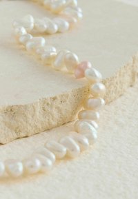Collier en perles d'eau douce avec des formes irrégulières ; principalement blanches avec une perle rose unique. Présenté sur une surface de pierre texturée.