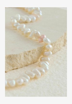 Freshwater pearl necklace with irregular shapes; predominantly white with a single pink pearl. Set against a textured stone surface.