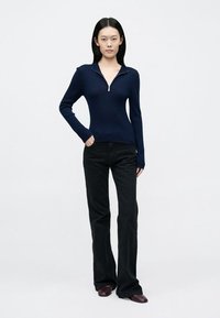 Tengerészkék bordázott kötött pulóver cipzáros magas nyakkal, fekete bővülő szárú farmerrel és sötétbarna cipővel párosítva. Egyszerű, testhezálló kialakítás.