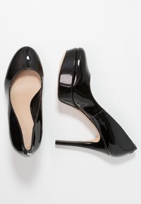 Escarpins hauts en cuir verni noir avec un bout arrondi élégant, un design découpé courbé et un talon aiguille fin. Texture lisse, style minimaliste.