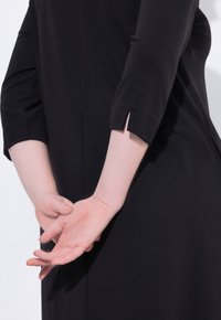 Schwarzes Kleid mit Dreiviertelärmeln, das ausgeschnittene Akzente an den Manschetten aufweist. Nahaufnahme von Händen, die hinter dem Rücken verschränkt sind. Glatte Stofftextur.