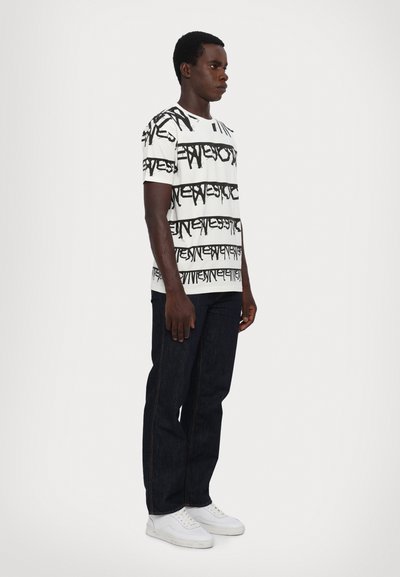Weißes T-Shirt mit schwarzem abstraktem Textmuster, kombiniert mit dunklen Jeans und weißen Sneakern. Das T-Shirt hat kurze Ärmel und einen Rundhalsausschnitt.