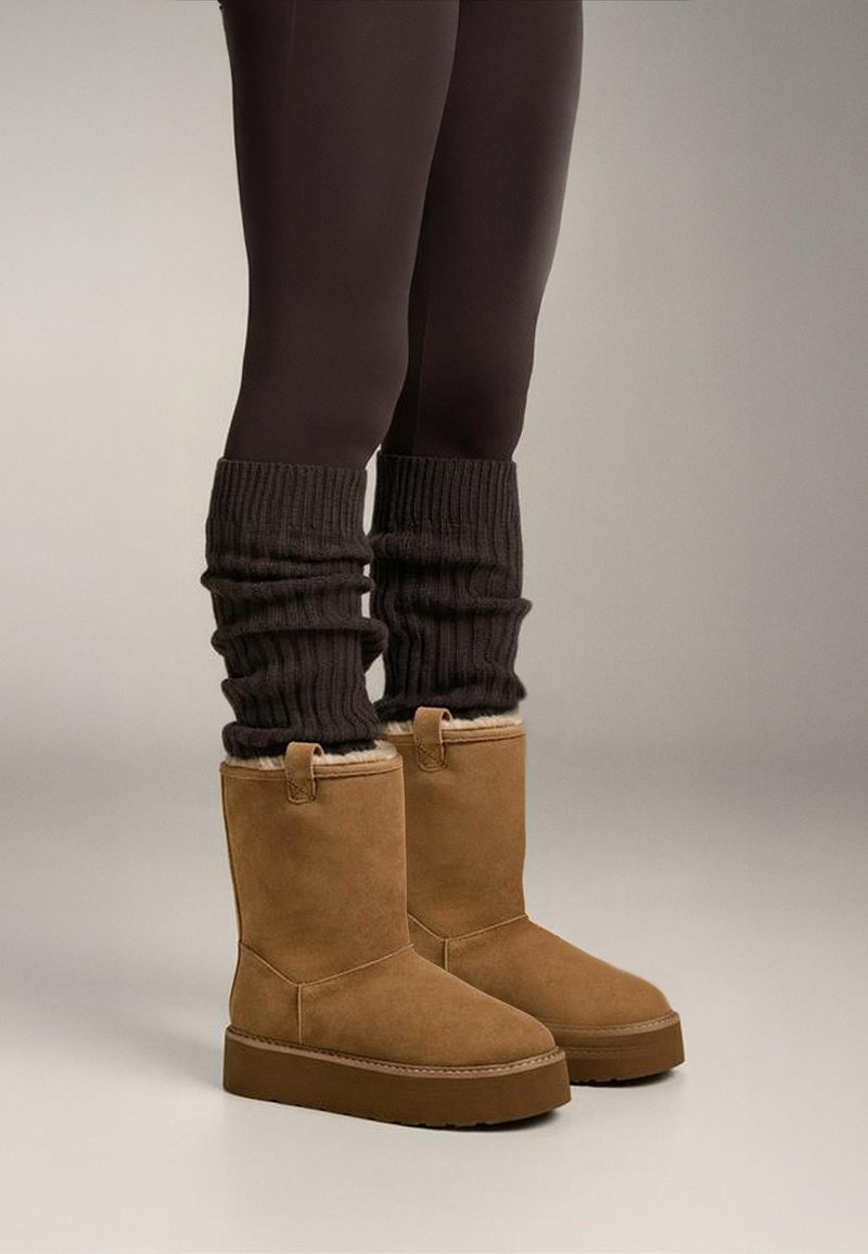 Brown suede ankle boots with a rounded toe and plush lining, featuring a flat sole and two side loops. Paired with dark ribbed leg warmers.