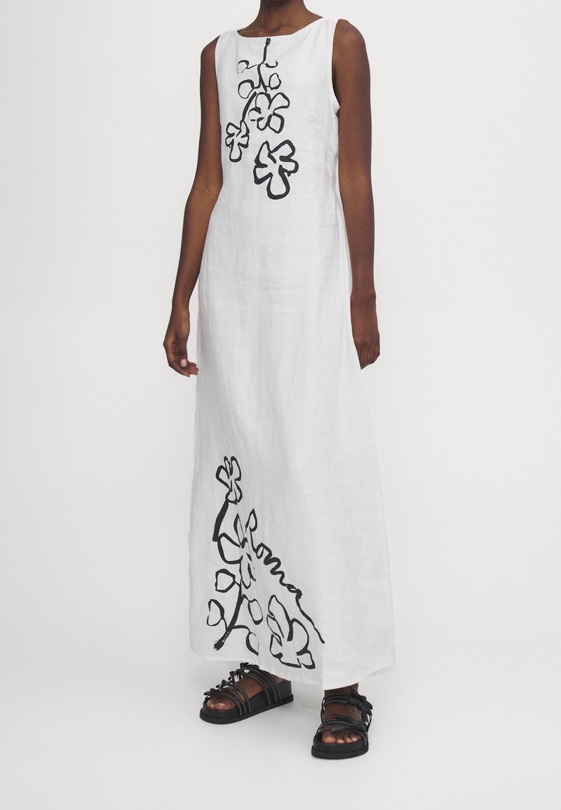 Valkoinen pellava-asu mustalla kukkakirjailulla, hihaton malli ja rento istuvuus. Omistaa pyöreän kaula-aukon ja lattiaan ulottuvan siluetin.