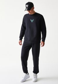 Μαύρο φούτερ με γραφικό ταύρου σε τυρκουάζ χρώμα, συνδυασμένο με μαύρη φόρμα και μαύρο καπέλο με λογότυπο ταύρου, φορεμένο με λευκά αθλητικά παπούτσια.