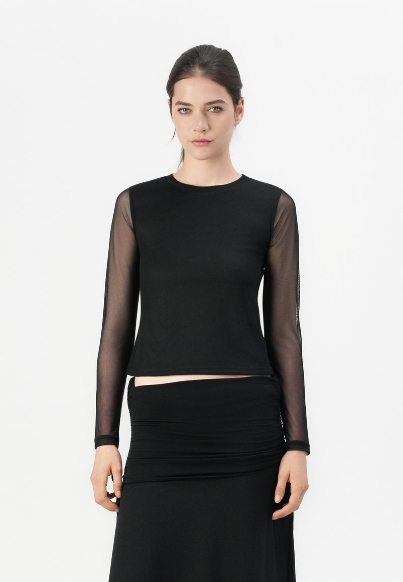 Black long-sleeve top with sheer mesh sleeves, round neckline, and fitted design. Paired with a black ruched skirt. Smooth texture.
