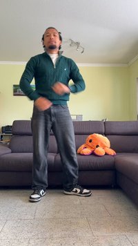 Persona vestida con un suéter de punto en verde oscuro y jeans oscuros, de pie sobre un suelo de baldosas, con un pulpo de peluche naranja en un sofá marrón.