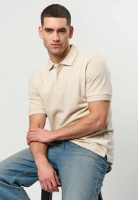 Jeune homme assis sur un tabouret portant un polo beige clair et un jean bleu, regardant droit devant avec une expression neutre.