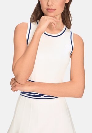 Woman in sleeveless white top with navy blue trim and matching skirt, arms crossed, hand near chin, neutral background.