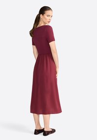 Robe bourgogne à manches courtes, corsage ajusté et jupe fluide. Portée avec des chaussures noires texturées comportant des accents décoratifs.