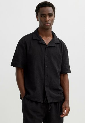 Hombre con cabello corto y rizado que lleva una camisa negra de manga corta y botones holgada y pantalones negros a juego, de pie con las manos en los bolsillos.