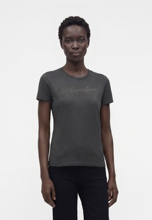 Femme aux cheveux courts et bouclés portant un t-shirt gris foncé ajusté avec un texte en cursive et un pantalon noir, debout devant un fond clair uni.