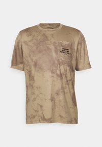 Béžové tričko s krátkým rukávem s texturovaným kamuflážním vzorem, které má také náprsní kapsu s černým písmem a kulatým výstřihem.