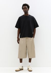 Črna oversized majica, bež plisirane kratke hlače, bele nogavice in teniske z rjavimi detajli, vse postavljeno na enobarvno ozadje.