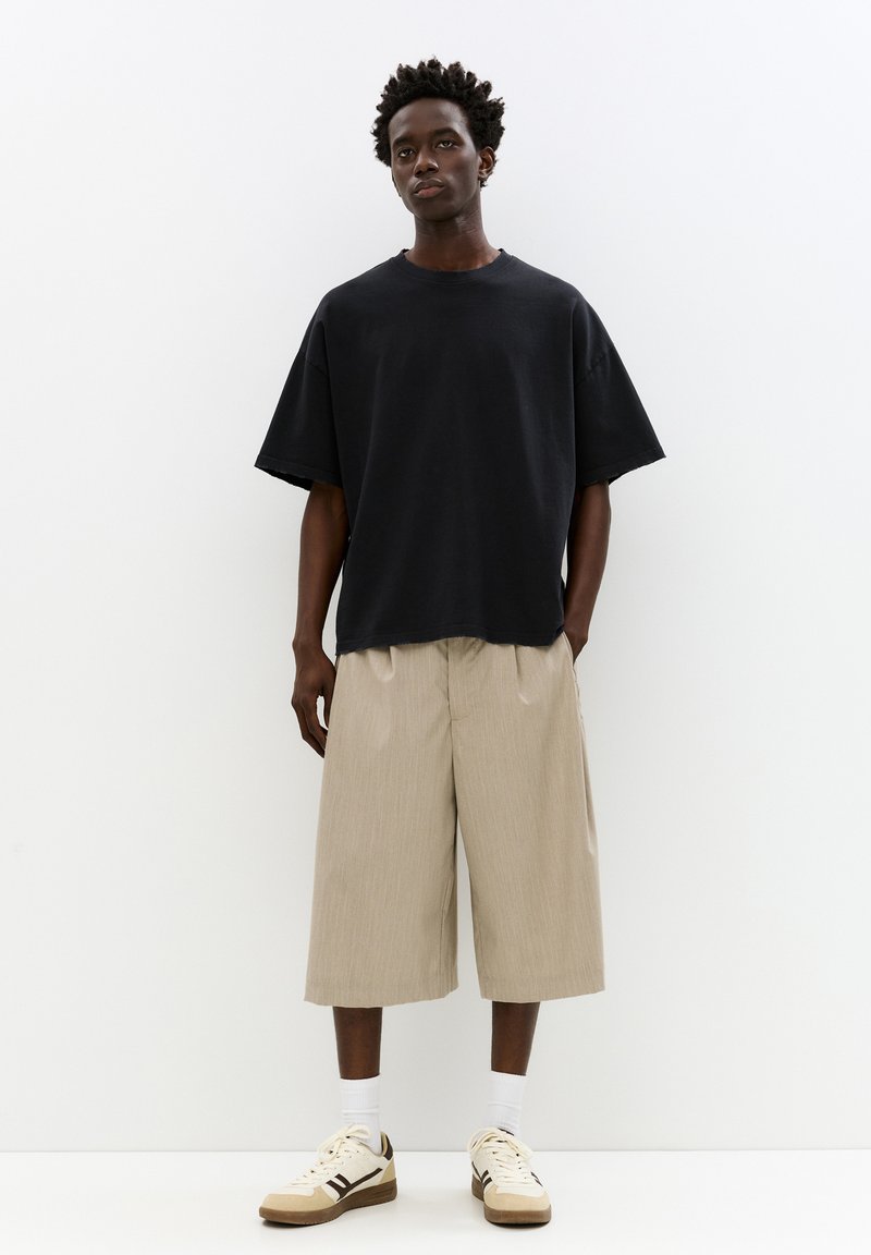 Črna oversized majica, bež plisirane kratke hlače, bele nogavice in teniske z rjavimi detajli, vse postavljeno na enobarvno ozadje.