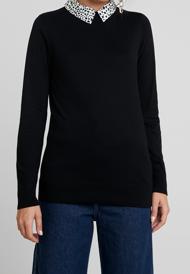 Femme portant un haut noir à manches longues avec un col tacheté blanc et noir, associé à un jean bleu sur fond neutre.
