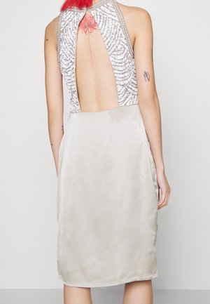 Osoba o rudych włosach w srebrnej, bezrękawnikowej bluzce z cekinami i otwartymi plecami oraz jasnej, satynowej spódnicy midi, ukazująca tatuaże na plecach.