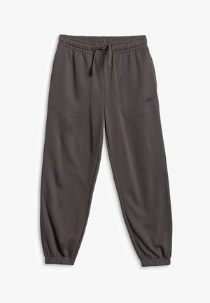 Garçon debout, bras croisés, portant une chemise blanche à manches courtes, un pantalon de jogging foncé et des baskets blanches sur fond gris.