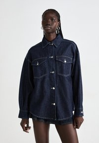 Dark blue denim jacket with a collar, long sleeves, two front pockets, and a button closure. Features contrast stitching and silver hardware accents.