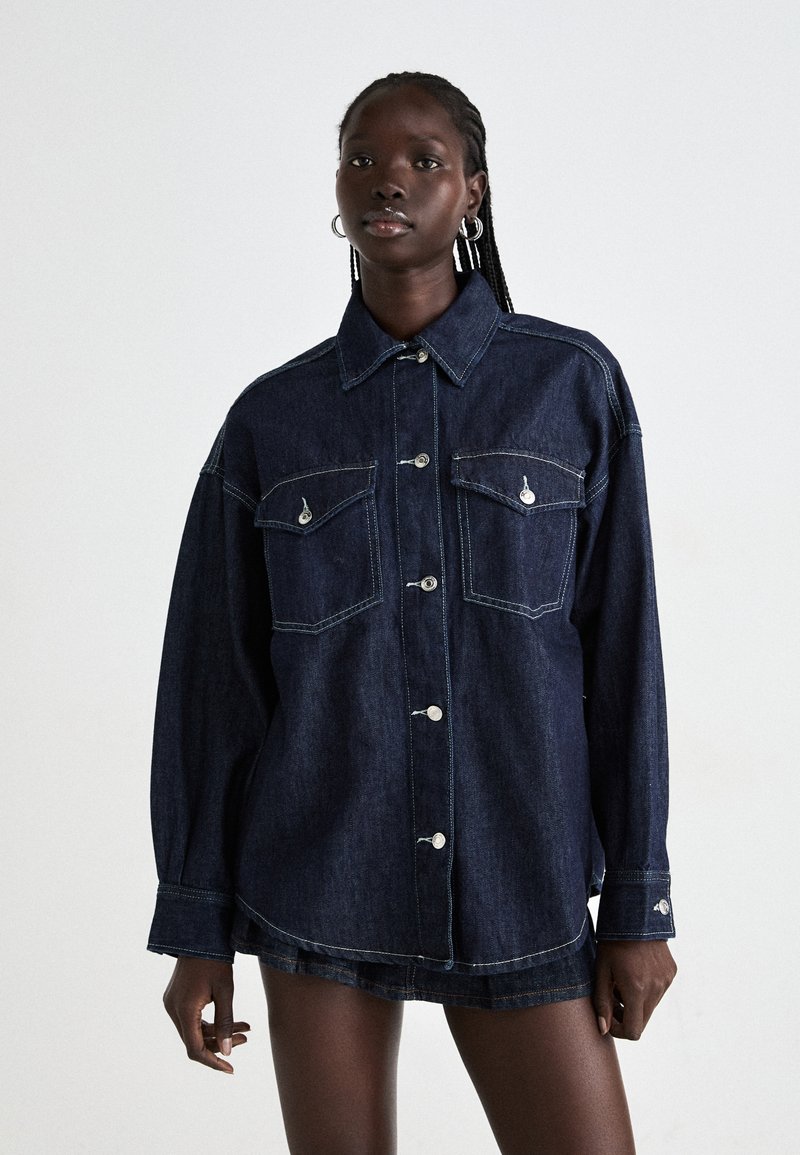 Dark blue denim jacket with a collar, long sleeves, two front pockets, and a button closure. Features contrast stitching and silver hardware accents.