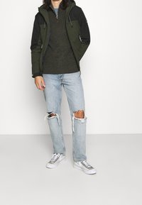 Pánská zeleno-černá bunda přes tmavě zelený pletený svetr, doplněná světle modrými roztrhanými džínami a bílými teniskami se stříbrnými detaily.