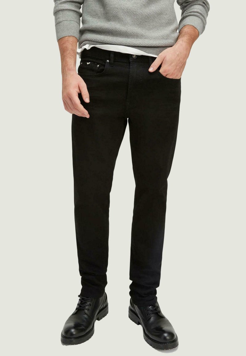 Jeans de mezclilla negros con un ajuste slim, que presentan un diseño clásico de cinco bolsillos, costuras sutiles y herrajes metálicos. Combinados con botas negras con cordones.