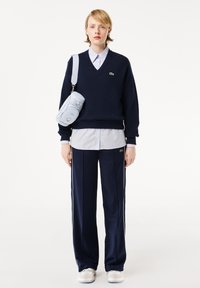 Marineblauwe v-hals trui, lichtblauw overhemd met knopen, marineblauwe trainingsbroek met witte strepen, en een kleine lichtblauwe crossbody tas. Witte sneakers.