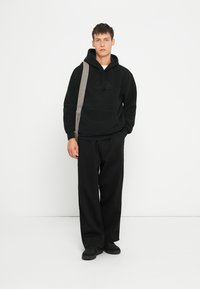 Felpa in pile nera con tasca frontale e logo tonale, abbinata a pantaloni neri larghi e scarpe nere. Cinghia regolabile a tracolla.
