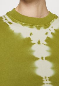 Zielony sweter tie-dye z zaokrąglonym, żebrowanym dekoltem oraz nieregularnym białym wzorem wtopionym w materiał, podkreślający teksturowane wykończenie.