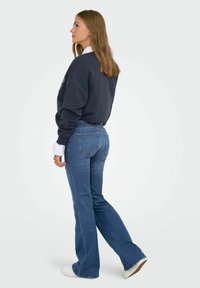 Tamno plavi sweatshirt opuštenog kroja, ispod njega bijela košulja s kragnom, i plave traperice s širom nogavicom. Model nosi bijele tenisice.