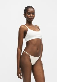 Γυναίκα με πλεξούδες, φορώντας λευκό κοντό τοπ και μπεζ σλιπ μπικίνι, στέκεται μπροστά σε απλό φωτεινό φόντο.