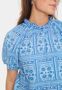 Blusa blu con motivo floreale, maniche a palloncino corte, scollatura arricciata e chiusura con bottoni, realizzata in tessuto leggero con una consistenza morbida.