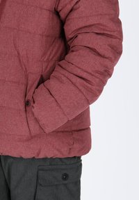 Giacca puffa rossa texturizzata con chiusura a zip e tasche laterali, caratterizzata da un design con polsini flessibili. Abbinata a pantaloni cargo grigi scuri.