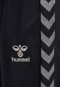 Pantalon de sport noir avec un motif en chevrons texturé argenté sur le côté. Présente un logo brodé en blanc de "hummel" près de la taille.