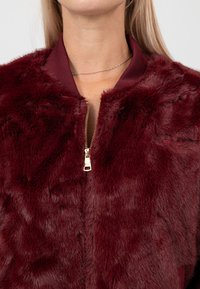 Giacca bomber in pelliccia finta bordeaux con zip dorata, collo a costine e una texture morbida. Presenta un accento ricamato discreto.