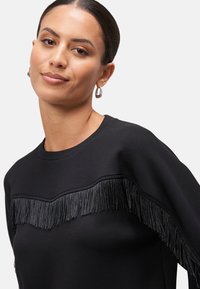 Černá mikina s texturovaným střapcem na hrudi, klasickým kulatým výstřihem a dlouhými rukávy. Hladký povrch látky.