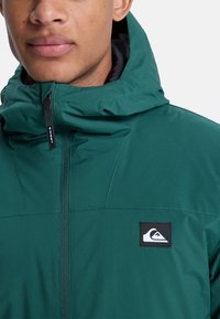 Groene hooded jacket met een rits aan de voorkant, voorzien van een zwart logo patch, een zachte textuur en naaddetails op de borst.