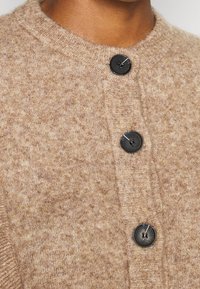 Gilet en tricot beige avec un col rond et trois boutons noirs. Il est fabriqué en tissu doux et texturé avec un motif subtil.